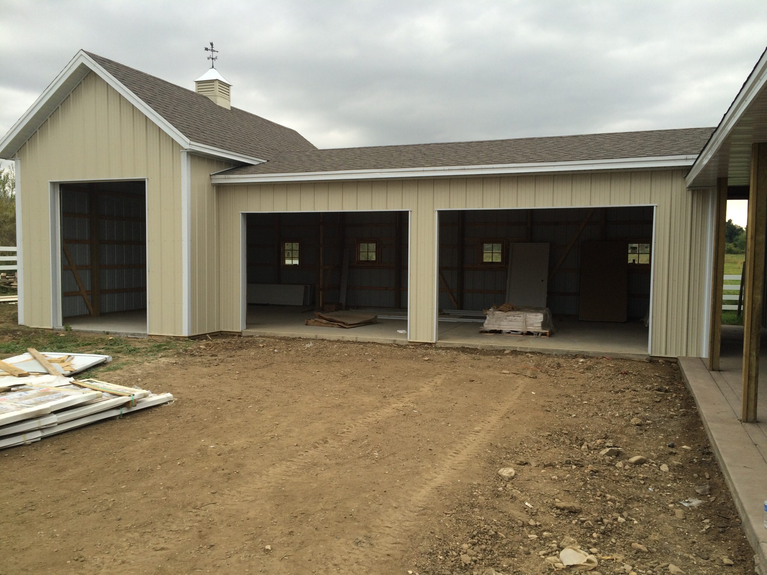 A beige metal garage or storage building with three open bays, unfinished interior, and dirt ground in front under a cloudy sky.
