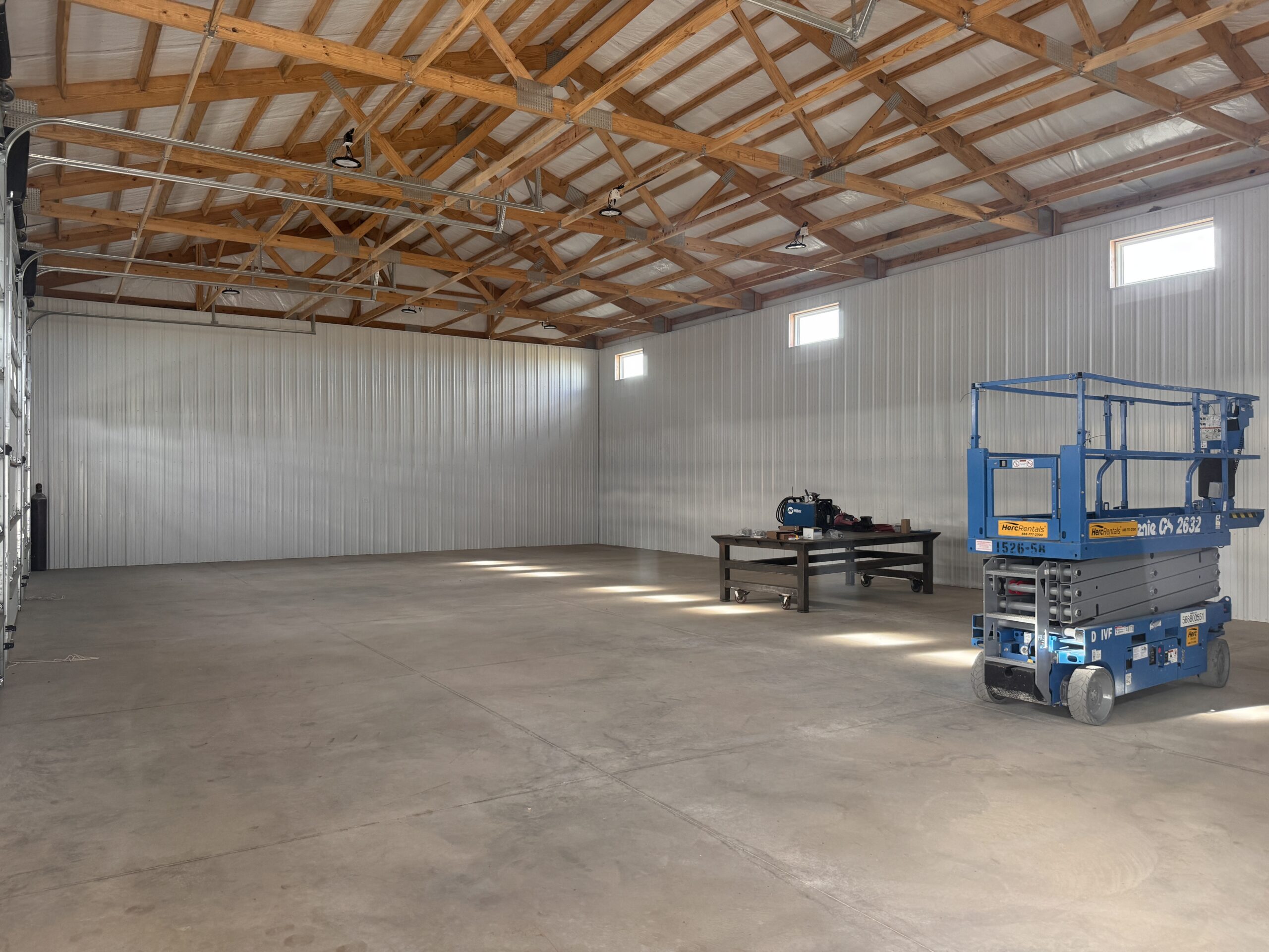 An empty metal building with a concrete floor, a metal workbench, and a blue scissor lift.