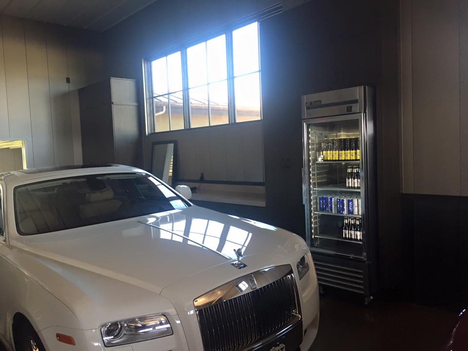 A white Rolls-Royce parked indoors next to a refrigerator stocked with cans of energy drinks and other beverages.