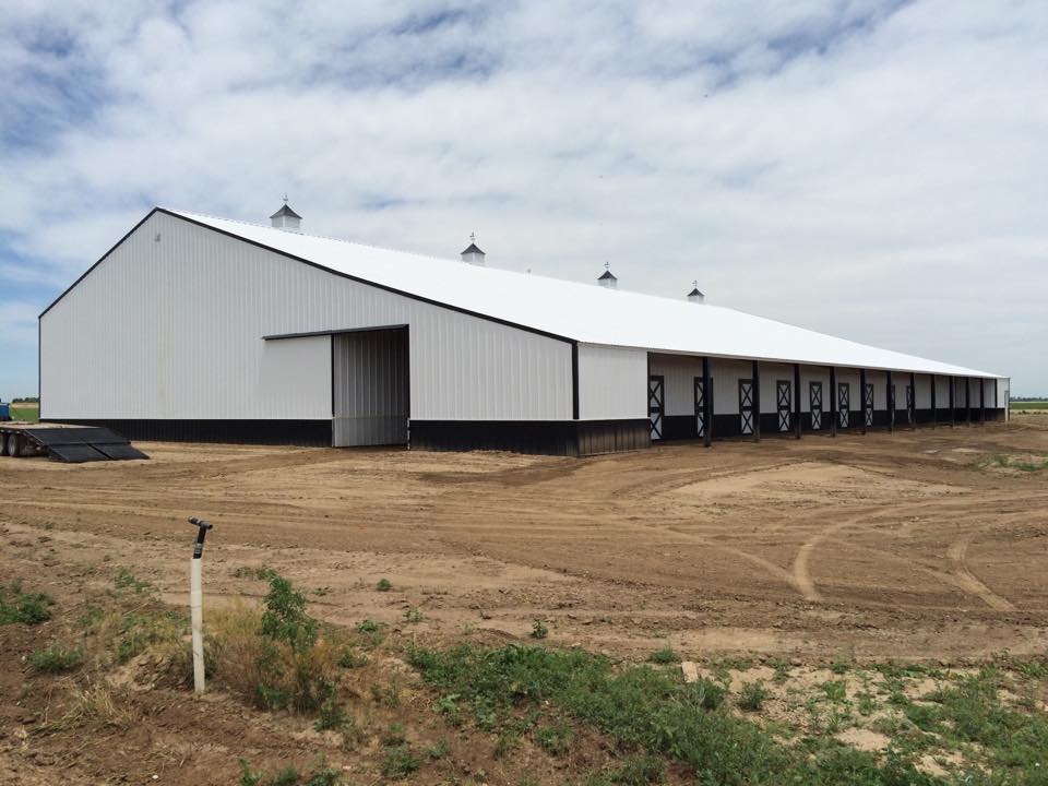 Large, white metal barn with multiple doors stands on a dirt lot under a partly cloudy sky, with some green vegetation in the foreground.