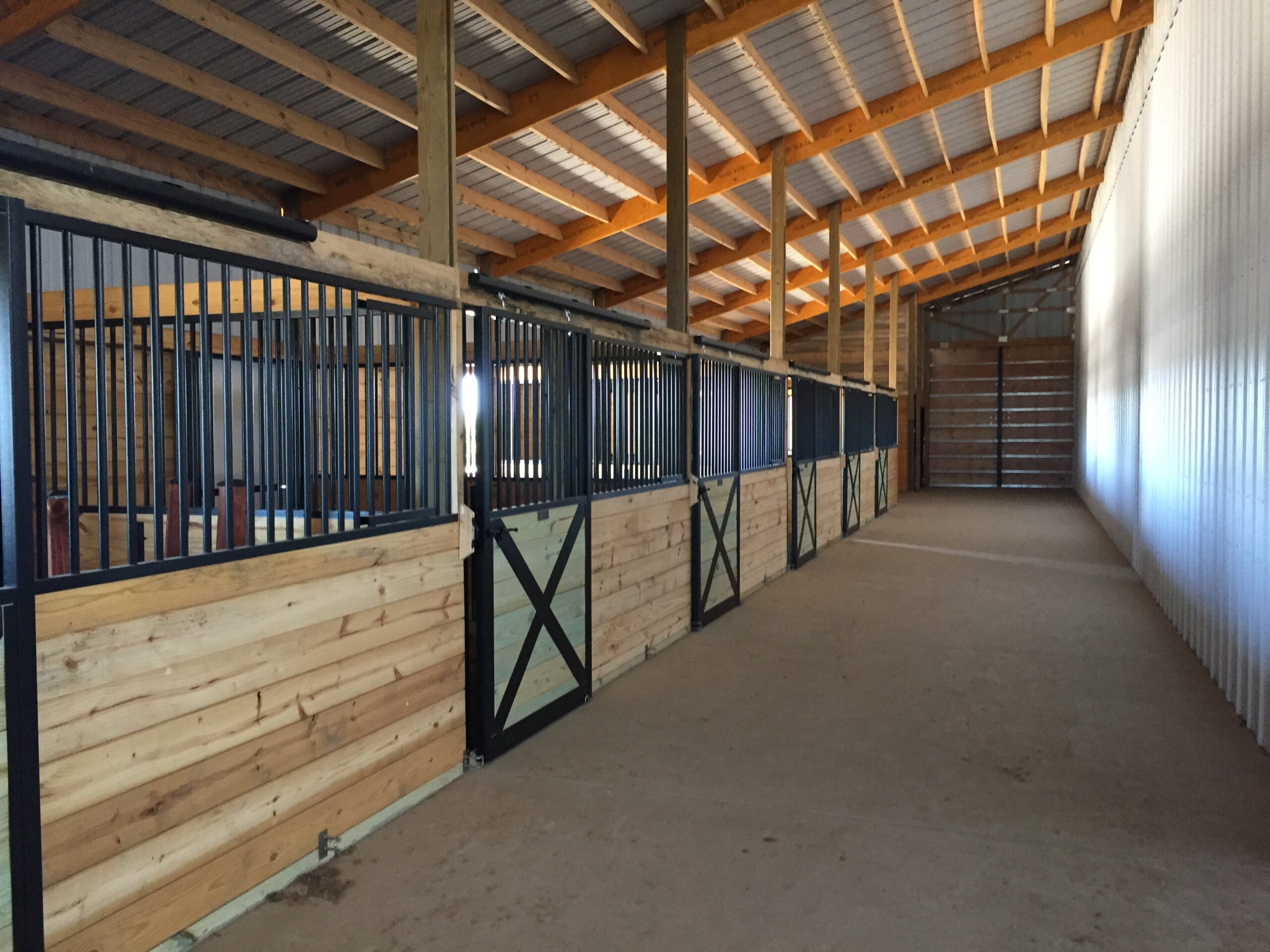 Interior of a clean, empty horse stable with wooden stalls and metal gates, concrete floor, and a metal roof with exposed beams.