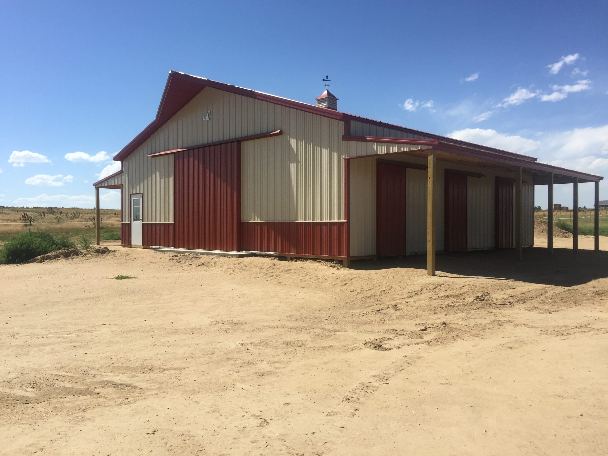 A large red and beige metal barn with a covered side area stands on a flat, sandy lot under a clear blue sky.