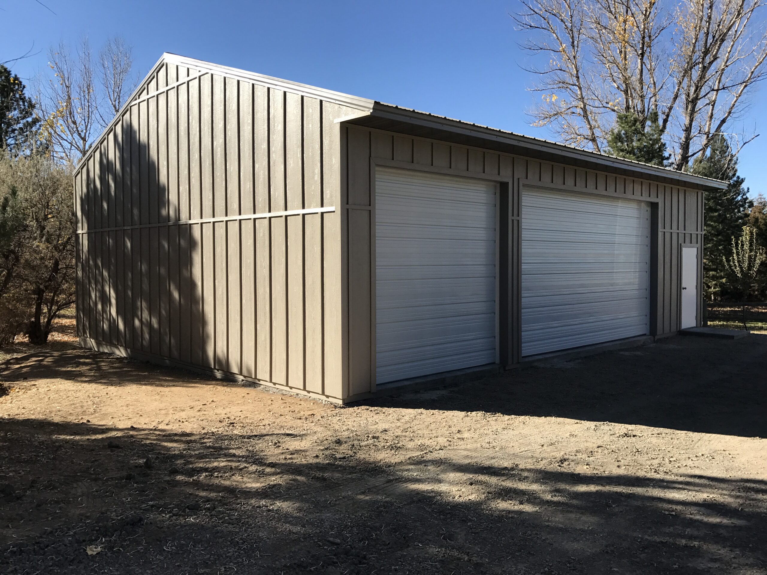 A large metal garage building with two closed white roll-up doors and a side entry door, surrounded by trees under a clear blue sky.