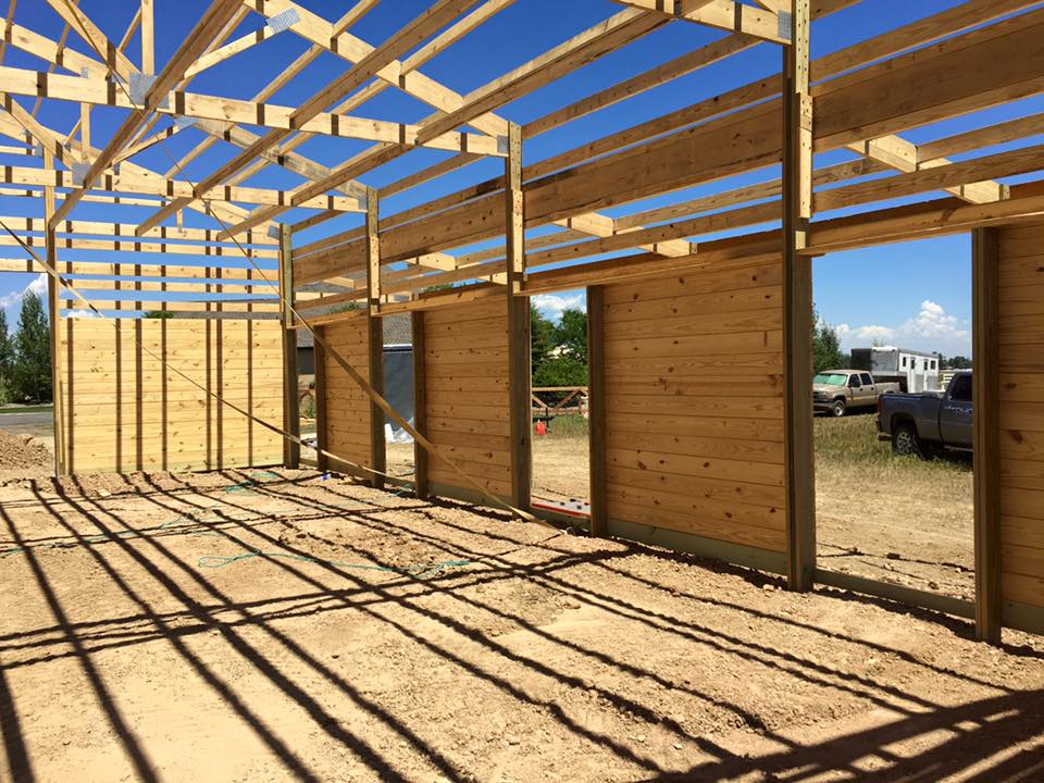 Wooden frame structure of a building under construction, with partial walls and exposed beams casting shadows on the dirt floor.